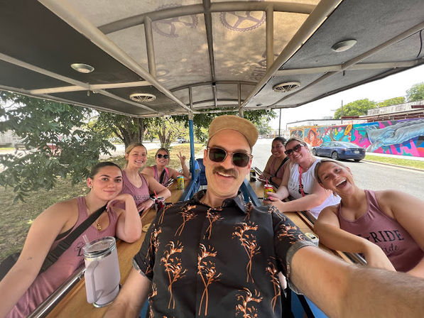 Group of friends on a pedal pub party bike taking a selfie on a sunny city street, man in hat and sunglasses in front, smiling women and a colorful mural along the roadside