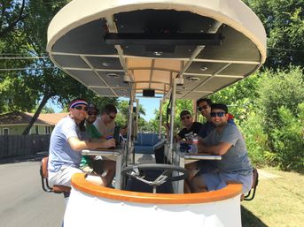 Six friends smiling and wearing sunglasses on a covered pedal‑pub party bike with a counter and drinks, rolling down a sunny suburban street lined with trees and houses.