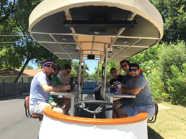 Six friends wearing sunglasses and caps sit at a bar-style pedal pub under a canopy, drinking and smiling while parked on a sunny suburban street lined with trees and houses.