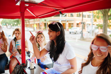 Friends laughing on a red pedal pub cart on a downtown street, a woman pointing excitedly while others in white shirts wear novelty sunglasses