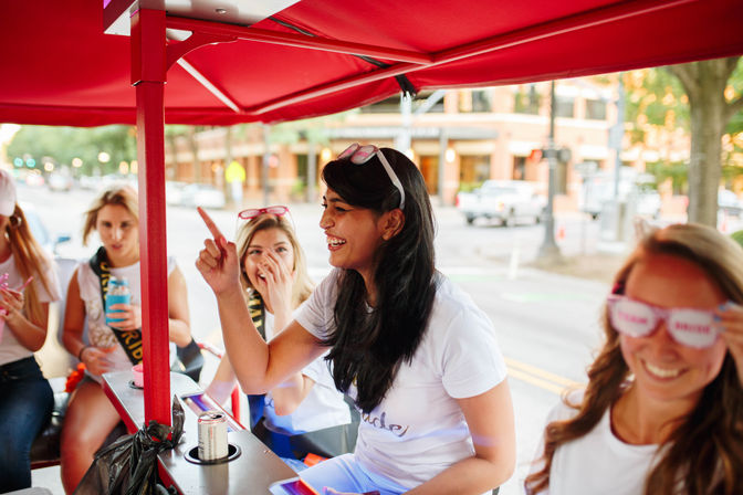 Group of women laughing on a red pedal-party bike under a canopy on a downtown street — bachelorette party vibe with sunglasses, drinks, and urban buildings in the background.