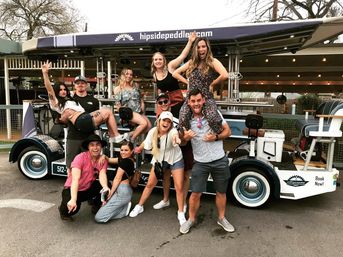 Ten friends laughing and posing around a pedal-powered party bike outdoors — one woman on a man's shoulders, bar-style seats and string lights visible.