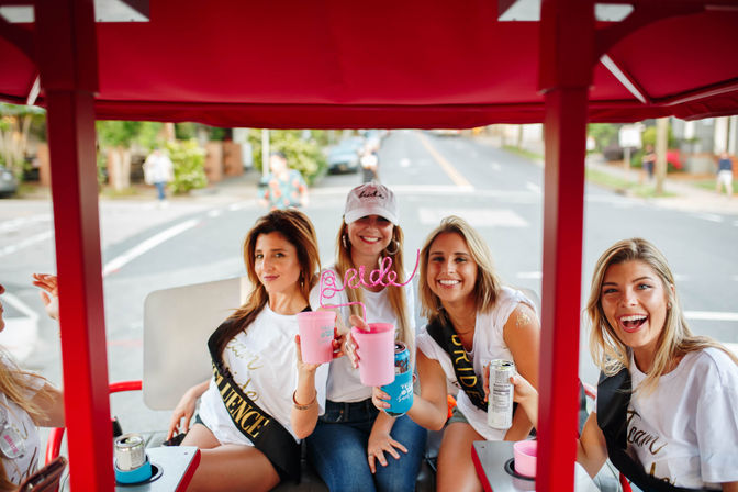 Bachelorette party of four friends smiling on a red pedal-powered party vehicle on a city street, wearing sashes and holding pink cups, canned drinks and a curly “bride” straw — outdoor urban celebration.