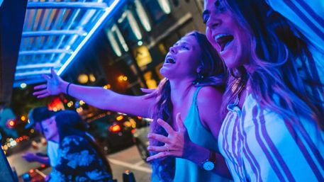 Two women singing and cheering under vibrant blue neon lights on a lively downtown street at night, with blurred city traffic and pedestrians in the background.