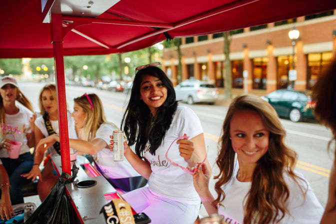 Smiling young women in white shirts riding a red-covered party bike on a downtown street, one holding a canned drink and pink sunglasses during a lively bachelorette-style outing.