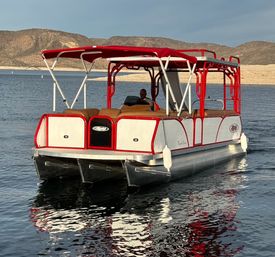 Sunlit red-and-white pontoon boat with canopy and tan seating gliding on a calm desert lake near arid, hilly shoreline, person at the helm.
