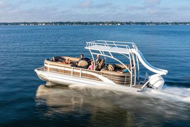 Pontoon boat with white waterslide and passengers speeding across a calm blue lake, leaving a foamy wake with a tree-lined shoreline and clear sky in the background.