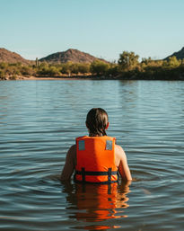 Sunlit swimmer in an orange life jacket wading in a calm lake, facing desert hills and scrubby shoreline under a clear blue sky.