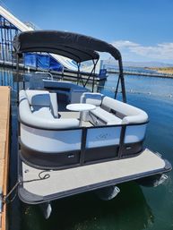 Pontoon boat docked at a sunny lake marina with a black canopy, light-gray wraparound seating and a small round table, water slide and distant shoreline under a clear blue sky