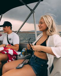 Two friends relaxing on a motorboat at a lake under a moody cloudy sky — young woman in a black bikini top, white shirt and denim shorts holding sunglasses, man in a cap with a red-striped towel and canned drinks in the cupholder, summer boating vibe.