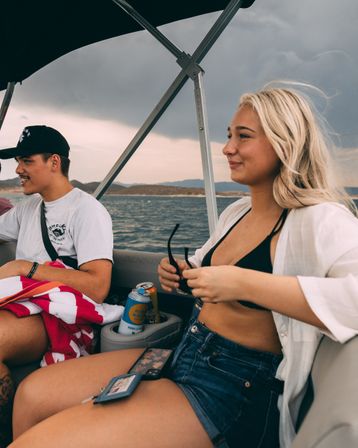 Two friends relaxing on a motorboat at a lake under a moody cloudy sky — young woman in a black bikini top, white shirt and denim shorts holding sunglasses, man in a cap with a red-striped towel and canned drinks in the cupholder, summer boating vibe.