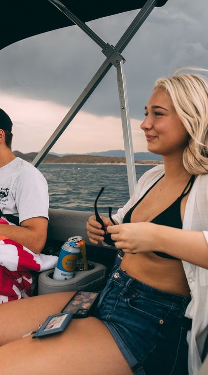 Two friends relaxing on a motorboat at a lake under a moody cloudy sky — young woman in a black bikini top, white shirt and denim shorts holding sunglasses, man in a cap with a red-striped towel and canned drinks in the cupholder, summer boating vibe.
