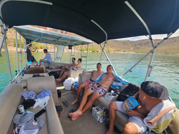 Friends relaxing on a covered pontoon boat over turquoise lake water with arid desert hills in the background, snacks and towels visible