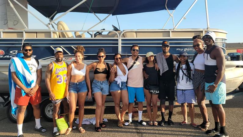 Group of friends posing in front of a pontoon boat in a sunny marina parking lot, wearing swimsuits, shorts and sunglasses for a fun summer day on the water