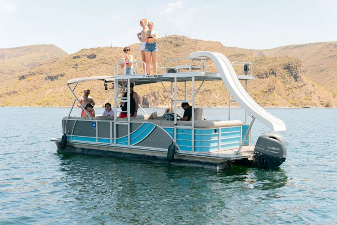 Pontoon boat with upper-deck water slide and a group of people enjoying a sunny summer lake day near dry, rocky hills