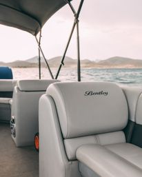 Light-gray pontoon boat seat with embroidered logo, overlooking a calm lake and rolling mountains under a soft sunset.