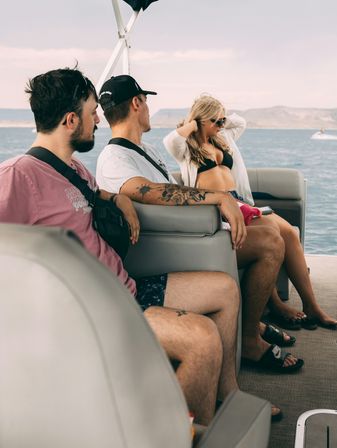 Three friends lounging on a pontoon boat over blue lake waters — woman in a black bikini and sunglasses adjusting her hair, a man with a floral arm tattoo and casual summer clothes.