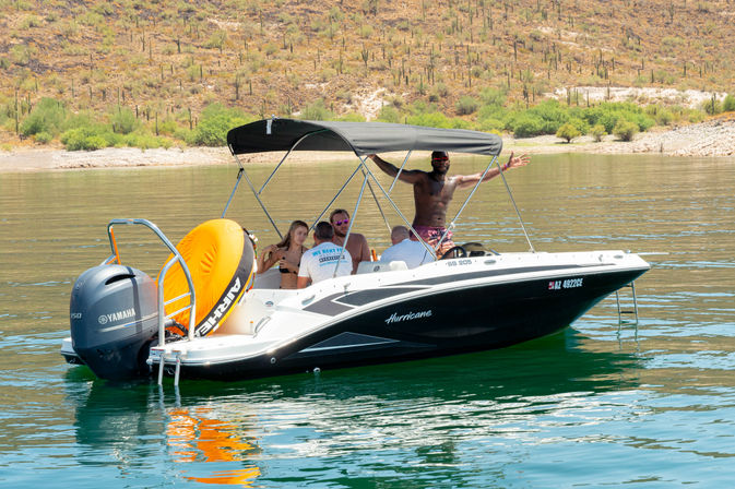 Group of friends on a black-and-white Hurricane boat at an Arizona desert lake — one person standing with arms wide, others seated under a black bimini with an orange towable tube and Yamaha outboard, calm green water and saguaro-covered hillside in the background.