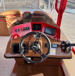 Sunlit pontoon boat helm with brown leather bench seats, glossy red dashboard, steering wheel, touchscreen display and throttle, docked at a lakeside wooden pier, ready to cruise.