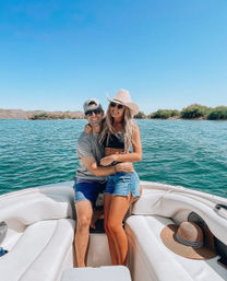 Smiling couple on a white boat enjoying a sunny summer day on a turquoise lake, woman in a wide-brimmed cowboy hat and denim shorts and man in a baseball cap and sunglasses with arid shoreline and low hills in the background.