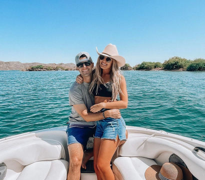 Smiling couple on a white boat enjoying a sunny summer day on a turquoise lake, woman in a wide-brimmed cowboy hat and denim shorts and man in a baseball cap and sunglasses with arid shoreline and low hills in the background.