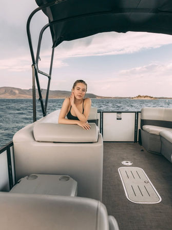 Person in a black swimsuit leaning on a seat aboard a pontoon boat on a lake with distant hills and a partly cloudy sky