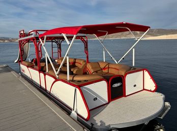 Bright red-and-white pontoon boat with red canopy and tan cushioned seats docked on a calm lake with sandy shoreline and low hills