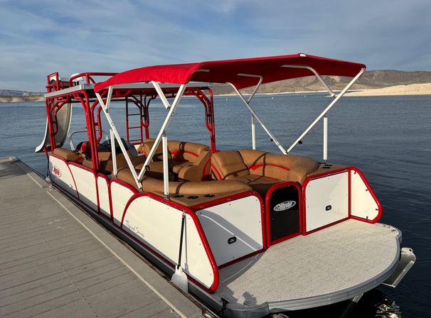 Bright red-and-white pontoon boat with red canopy and tan cushioned seats docked on a calm lake with sandy shoreline and low hills