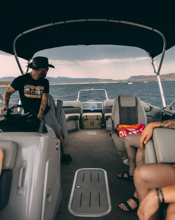 People relaxing on a pontoon boat cruising a mountain lake under an overcast sky; driver in a wide-brim hat at the helm, cushioned seats, insulated cooler and colorful towel.