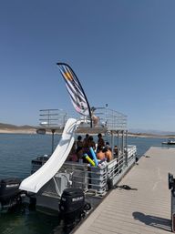 Pontoon party boat with a white water slide and colorful pool noodles moored at a sunny lake dock, swimmers in swimsuits on the deck and rolling hills on the distant shoreline.