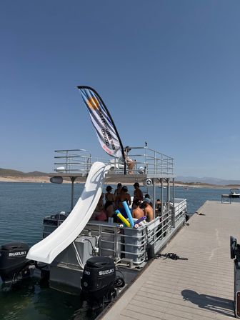 Pontoon party boat with a white water slide and colorful pool noodles moored at a sunny lake dock, swimmers in swimsuits on the deck and rolling hills on the distant shoreline.