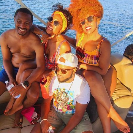 Sun-soaked group of four on a boat over blue water, wearing colorful swimwear and sunglasses and holding drinks and water bottles — lively summer lake outing.