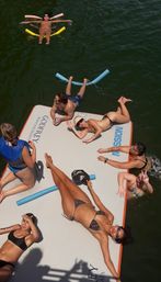 Overhead shot of a cheerful summer lake scene: friends sunbathing and lounging on a large floating platform in swimsuits while others float nearby with pool noodles and life vests.
