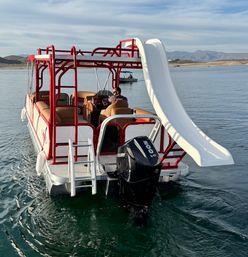 Red-and-white pontoon boat with a large white waterslide and Mercury V6 outboard on a calm mountain lake, passenger at the helm and distant shoreline and hills in the background.