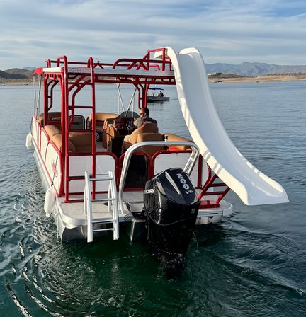 Red-and-white pontoon boat with a large white waterslide and Mercury V6 outboard on a calm mountain lake, passenger at the helm and distant shoreline and hills in the background.