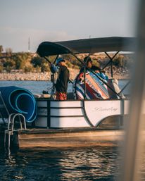 Pontoon boat on a lake at golden hour with two people prepping a colorful towable tube and ropes for watersports