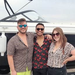 Three smiling friends in sunglasses and bold patterned shirts posing beside a white boat at a marina, summery casual vibe.