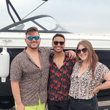 Three smiling friends in sunglasses and bold patterned shirts posing beside a white boat at a marina, summery casual vibe.