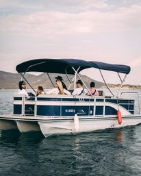 Friends lounging on a blue-and-white pontoon boat with a navy canopy, cruising a calm mountain lake with rolling hills and a soft cloudy sky.