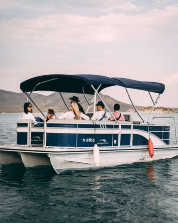 Friends lounging on a blue-and-white pontoon boat with a navy canopy, cruising a calm mountain lake with rolling hills and a soft cloudy sky.