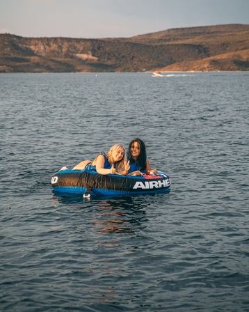 Two friends on a blue inflatable tube giving thumbs up while floating on a calm lake with hilly shoreline and a distant motorboat at sunset