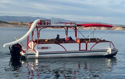 Red-and-white pontoon boat with waterslide and red canopy, person at the helm, cruising on a calm mountain lake