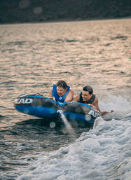 Two people in life jackets riding a blue inflatable tow tube across a lake, gripping handles as a boat's wake sprays water