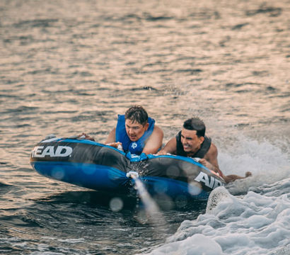 Two people in life jackets riding a blue inflatable tow tube across a lake, gripping handles as a boat's wake sprays water
