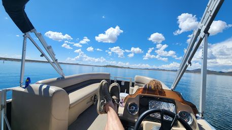 Relaxing view from a pontoon boat on a calm lake: legs propped up toward the helm, someone at the console, bright blue sky dotted with fluffy clouds and distant shoreline hills.