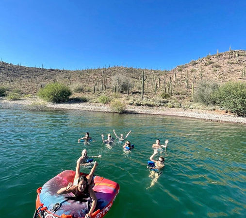People swimming and lounging on a colorful inflatable tube in a clear desert lake with saguaro cacti and rocky shoreline beneath a bright blue Arizona/Sonoran Desert sky.