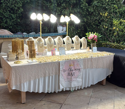 Outdoor patio jewelry display: lace-covered table with illuminated busts showcasing gold necklaces and bracelets, bright lamps overhead, vase of pink flowers and lush green hedge backdrop.