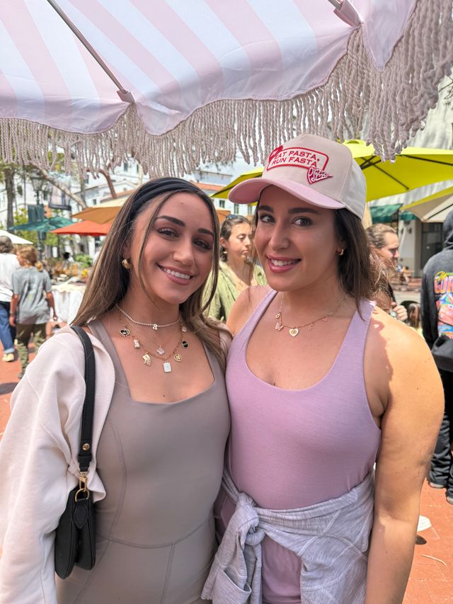 Two smiling women posing under a pink-and-white fringed umbrella at a sunny outdoor market with colorful patio umbrellas in the background