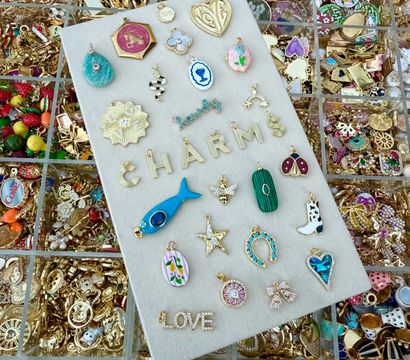 Colorful jewelry display of assorted gold and enamel charms and tiny pendants — blue fish, horseshoe, star, heart, floral medallions and letter charms spelling CHARMS and LOVE arranged on a cream board over divided trays of loose gold charms.