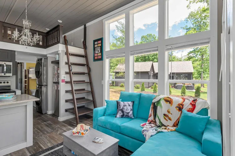 Bright modern cottage living room with turquoise sectional, pop-art face throw pillow, loft ladder, open kitchen island and a wall of tall windows overlooking a leafy backyard